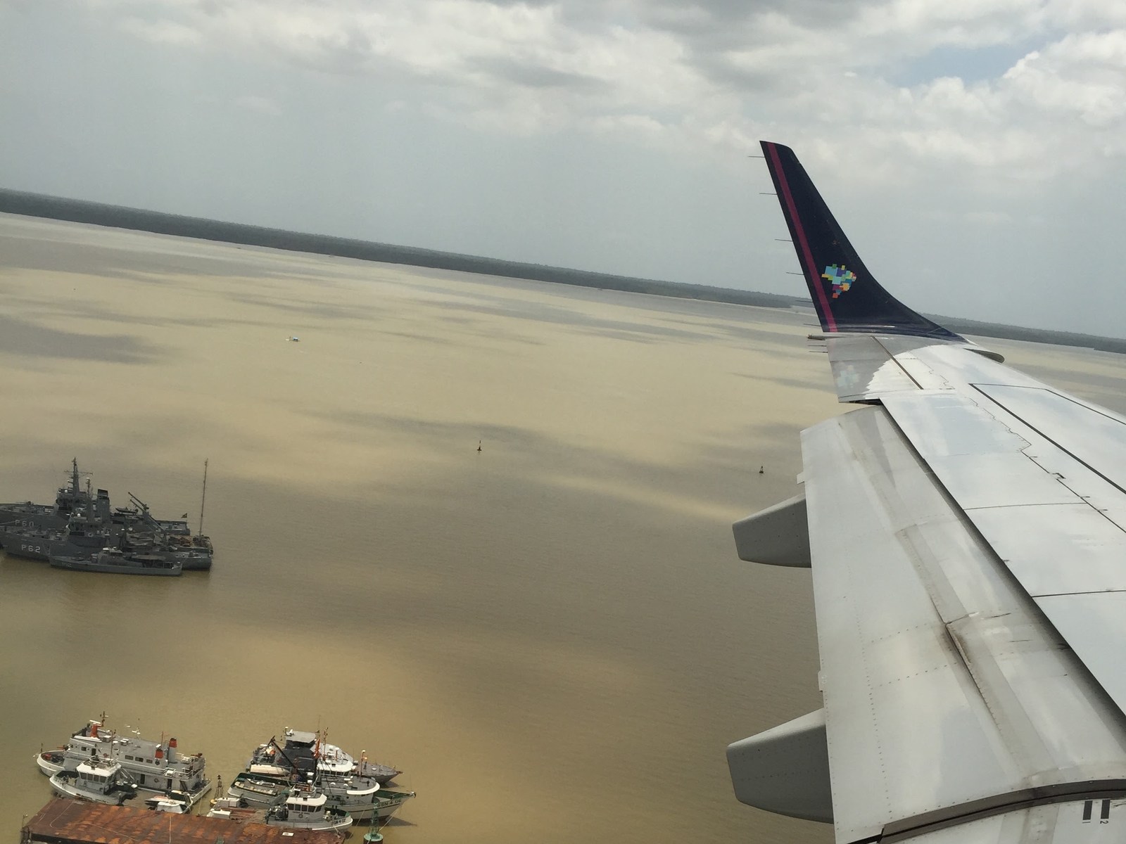 Vista da janela do avião sobre o rio, com a asa à direita e embarcações abaixo